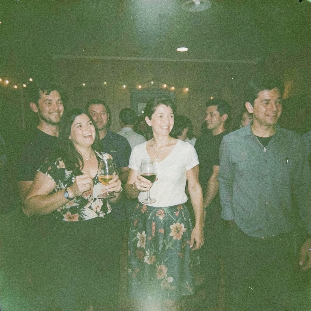 A grainy film photo of a couple at a reception, the groom raising a glass with an exaggerated grin while the bride rolls her eyes playfully; soft candlelit glow and muted earth tones.