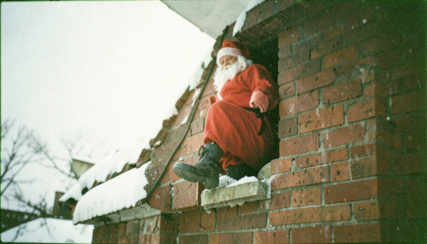 Grainy film photo of a Santa impersonator stuck halfway in a chimney with only his boots and red pants visible against a snowy rooftop, muted whites and brick reds.